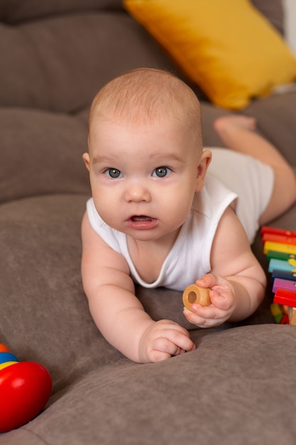 Cute toddler in white bodysuit lies at home on grey sofa with yellow pillows playing with wooden developing toy