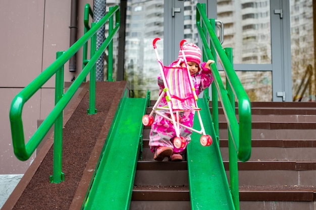 Cute toddler with a toy stroller walks along steel railing ramp for wheelchair carts and strollers