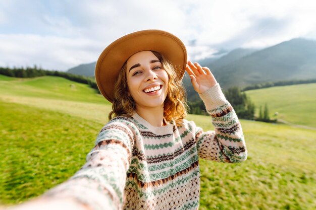 Cute traveler in hat takes selfie against backdrop of green mountain landscapes Concept of freedom