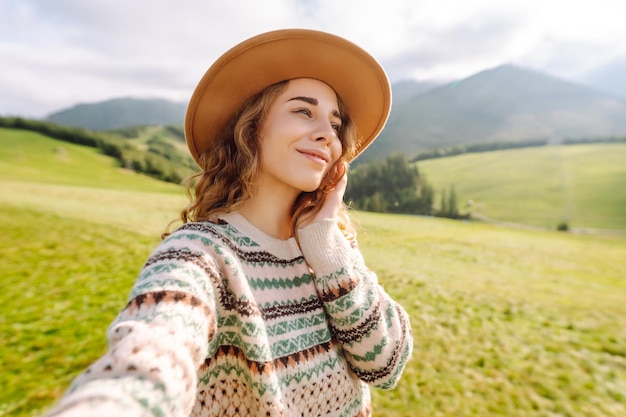 Cute traveler in hat takes selfie against backdrop of green mountain landscapes Concept of freedom