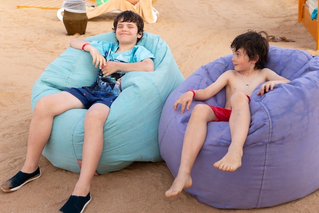 Cute two boys are sitting on the beach bean bag multicolored chairs at relaxing zone in a tropical resort.