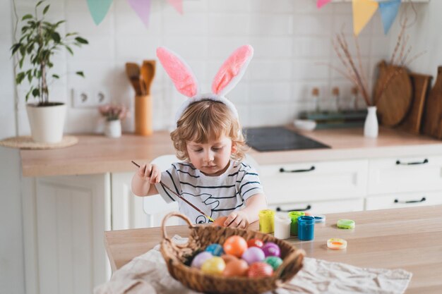 Cute twoyearold boy in bunny ears paint Easter eggs with multicolor paints sitting at the table on the kitchen