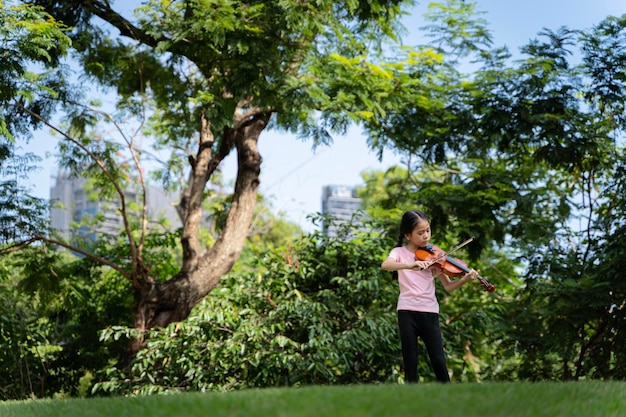 A cute white Asian girl is playing the violin on a grassy hill amid beautiful nature greenery and sunlight She is learning about new things that interest her