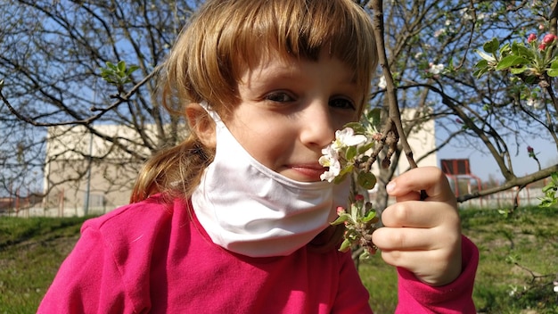 Cute white girl of 6 years old takes off a white protective mask and sniff beautiful fragrant flower