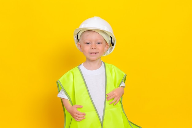 A cute white little boy, dressed in a reflective vest and a white hard hat, stands on a yellow wall.