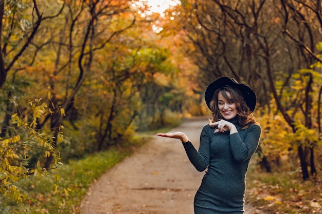 Cute woman in a black hat and a dark dress stands in the park in the autumn on the background of path