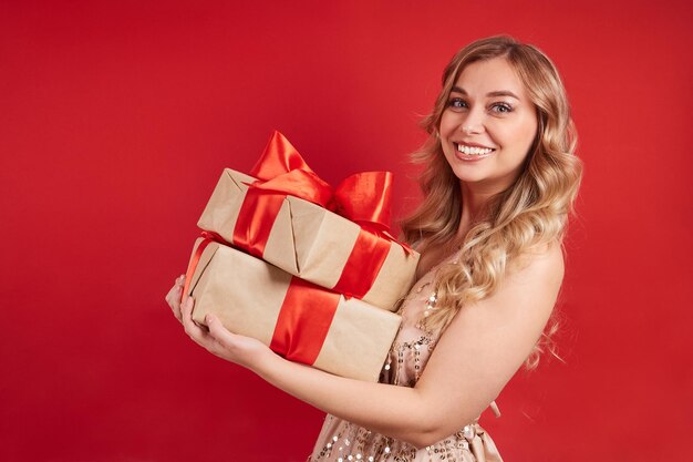 Cute woman in a dress holding gifts with her hands smiling looking at the camera on a red background