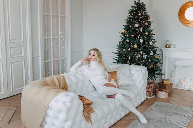 A cute woman in a dress is resting while sitting on a white sofa near a Christmas tree in a light interior of a cozy home