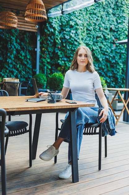A cute woman drinks coffee on the terrace of a summer cafe and works on a laptop remote work while on vacation in a cafe with a laptop