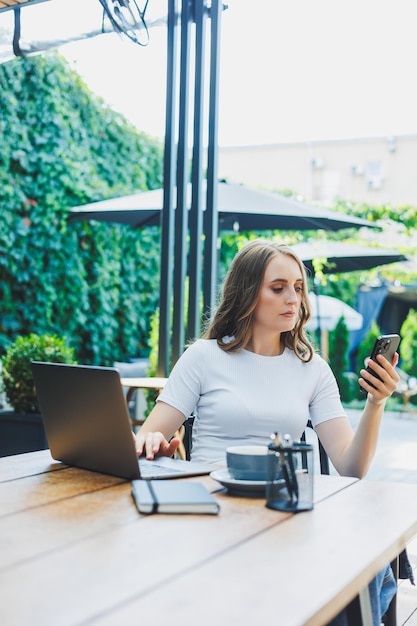 A cute woman drinks coffee on the terrace of a summer cafe and works on a laptop remote work while on vacation in a cafe with a laptop