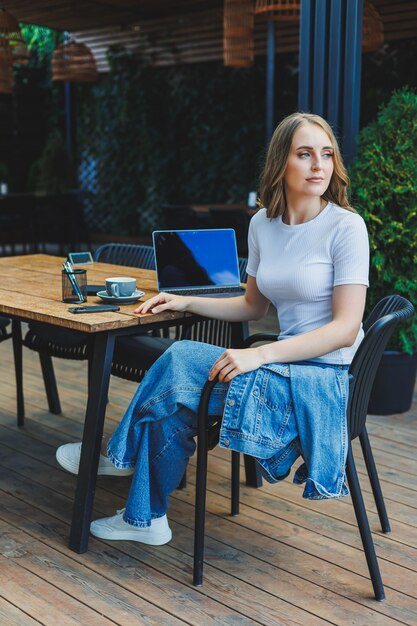 A cute woman drinks coffee on the terrace of a summer cafe and works on a laptop remote work while on vacation in a cafe with a laptop