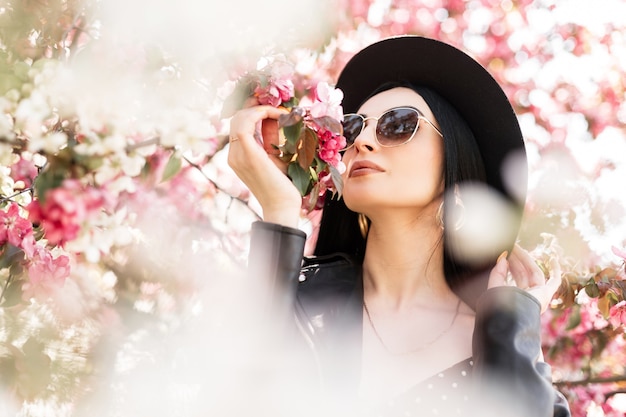 Cute woman in flowered park walks on nature. Lovely girl in elegant hat in trendy sunglasses in black fashionable outfit and pink spring flowers outdoors. Beauty lady sniffs amazing flowering tree.