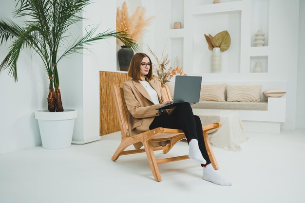 Милая женщина в очках и куртке сидит на стуле и работает на ноутбуке из гостиной удаленная работа из дома онлайн женщина-менеджер работает дома