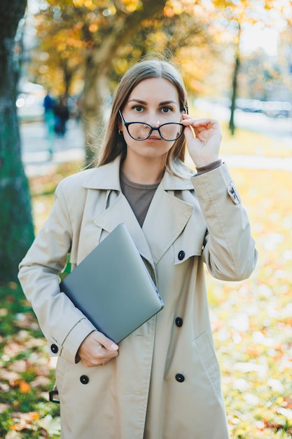 A cute woman in glasses with long hair is walking in an autumn park with a laptop in her hands Work online in the fresh air