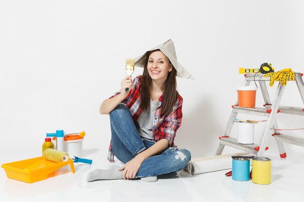 Cute woman in newspaper hat sitting on floor with brush, instruments for renovation apartment room isolated on white background. Wallpaper, accessories for gluing, painting tools. Repair home concept.