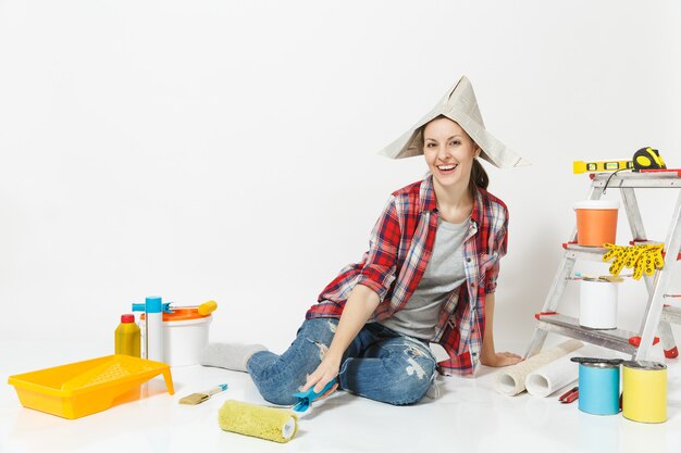 Cute woman in newspaper hat sitting on floor with paint roller, instruments for renovation apartment isolated on white background. Wallpaper accessories for gluing, painting tools. Repair home concept