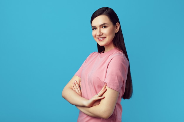 Cute woman in pink tshirt smiling and standing with hands crossed on blue