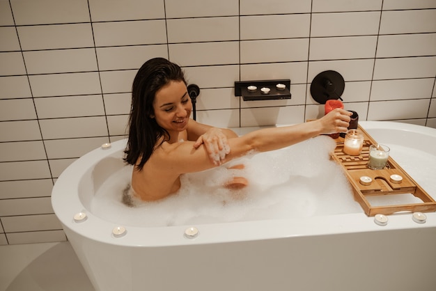 Cute woman relaxing in the beautiful bath full of foam decorated with candles. Healthy treatment and enjoyment