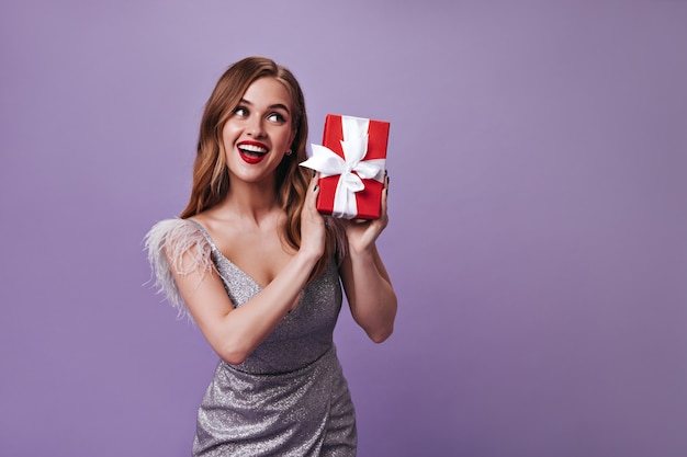 Cute woman in silver dress holding gift on purple wall