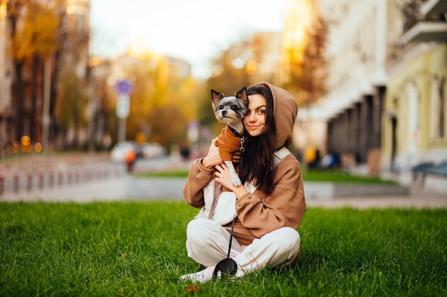 カメラを見ている彼女の腕の中でかわいい小さな犬と一緒に芝生の上に屋外で座っているかわいい女性