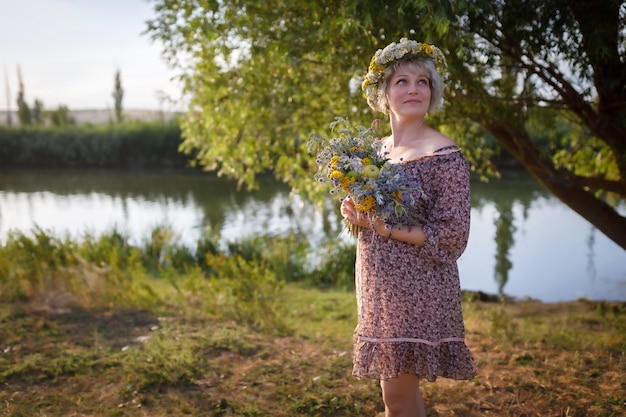 Милая женщина стоит на берегу реки с букетом полевых цветов