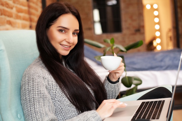 Милая женщина остается дома и работает на ноутбуке, пить кофе