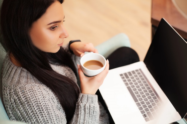 かわいい女性が家に滞在し、コーヒーを飲みながらラップトップで働く