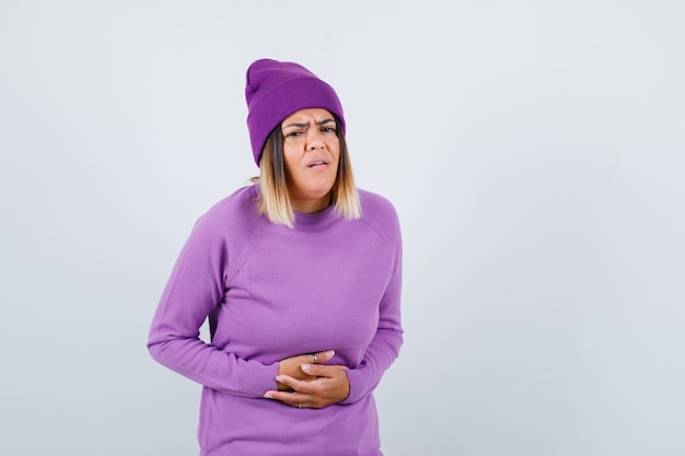 Cute woman in sweater, beanie suffering from stomach pain and looking unwell , front view.
