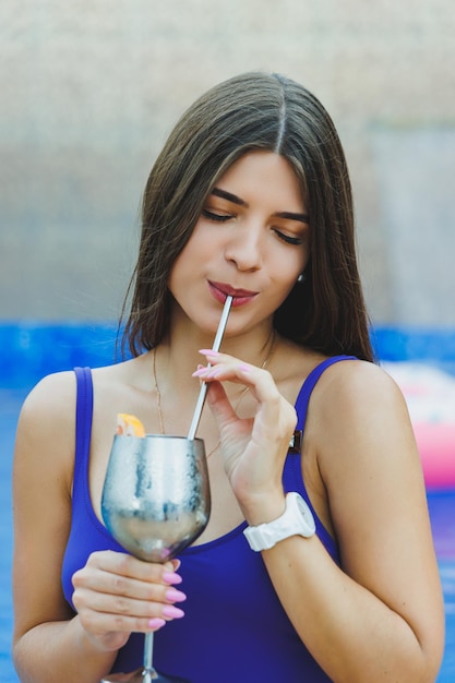 Cute woman in a swimsuit sitting by the pool and drinking a delicious cocktail and sunbathing by the pool during summer vacation