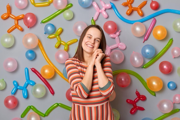 Cute woman with brown hair wearing striped dress looking at camera with charming smile expressing happiness standing against gray wall decorated with colorful balloons