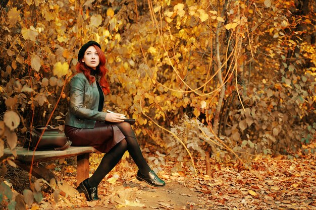 公園redhaiの公園秋の肖像画の木製ベンチに座っているグラフィックタブレットを持つかわいい女性...
