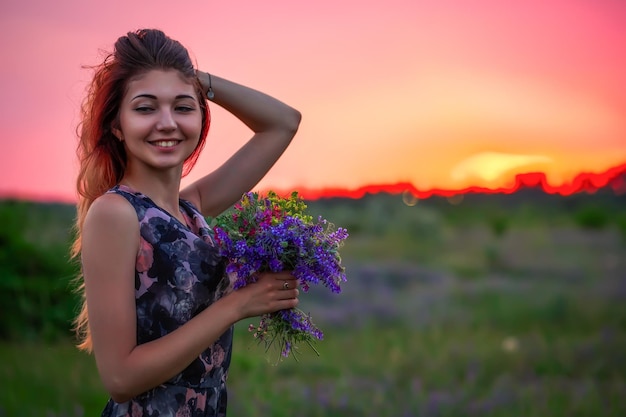 Cute young attractive girl with a bouquet of colorful flowers in her hands Evening walk in nature during sunset Pensive look Romantic atmosphere