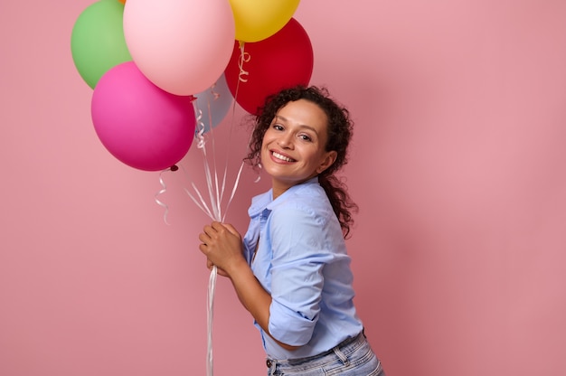 Cute young beautiful woman of mixed race ethnicity holding a lot of multicolored bright air balloons isolated over pink background with copy space for text and advertising