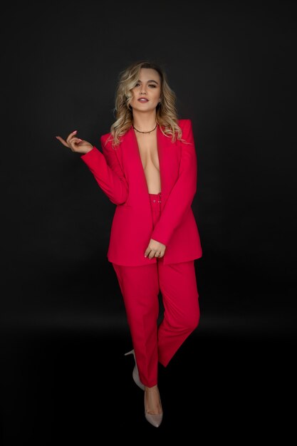 A Cute young blonde woman, smiling, looking at camera, posing in bright red pantsuit on black background in the studio.