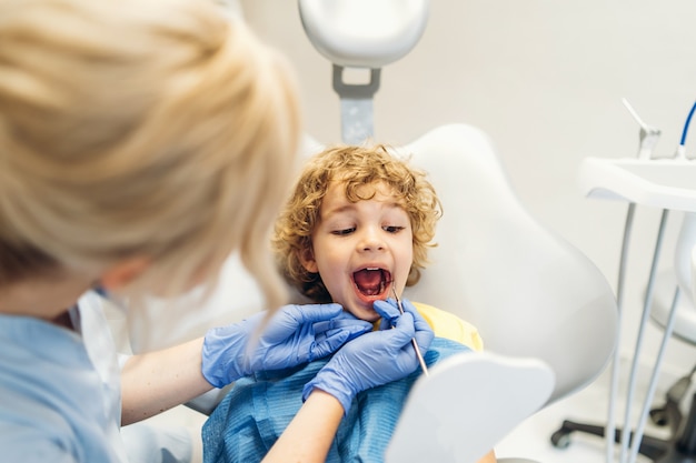 Cute young boy visiting dentist, having his teeth checked by female dentist in dental office.