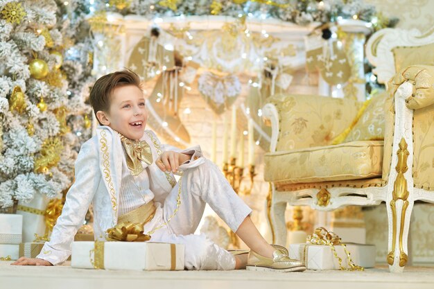 Cute young boy in white suit posing in room decorated to Christmas holiday