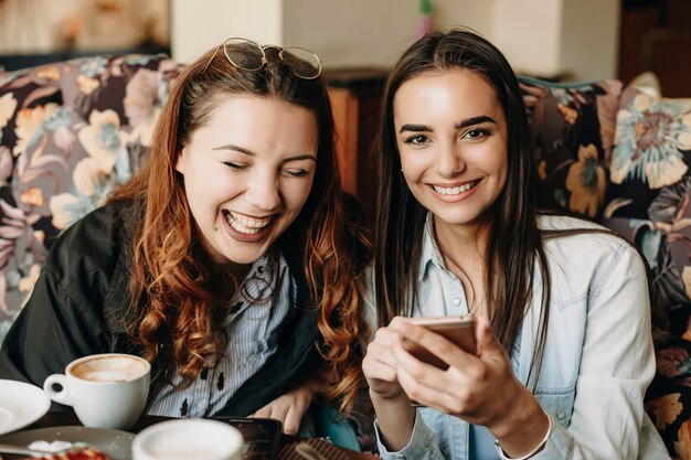 Cute young brunette looking at camera laughing while holding a smartphone while her girlfriend is laughing with closed eyes in a cafe.