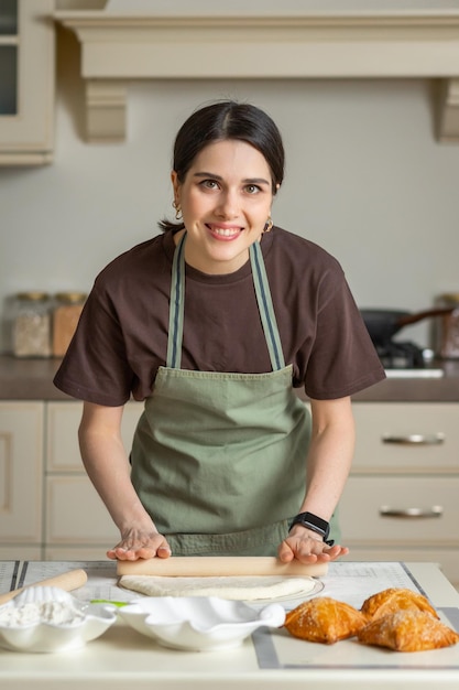 La giovane donna sorridente castana sveglia in un grembiule da cucina sta cucinando la cottura fatta in casa in cucina