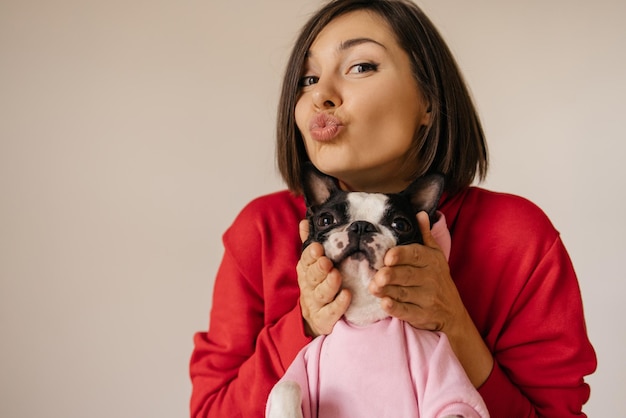 Cute young caucasian brunette woman touching cheeks of her french bulldog breed dog on white background Showing affection concept