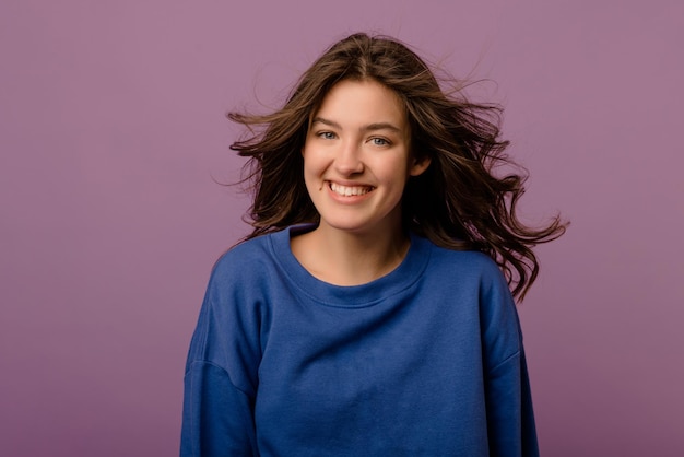 Cute young caucasian girl with flying dark hair in blue hoodie looks at camera on purple background Lifestyle female beauty concept