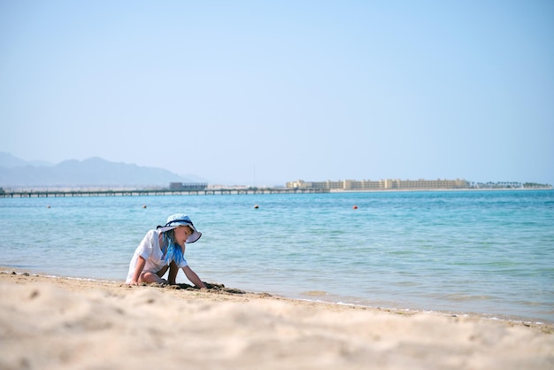 Foto cute bambina che gioca con la sabbia bianca sulla spiaggia del resort sullo sfondo del cielo azzurro, acqua limpida e riva lontana concetto di turismo e ricreazione estiva