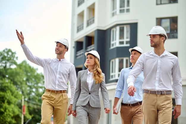 Cute young construction worker pointing at something to an attractive stylish blonde female supervisor and two male colleagues in hardhats