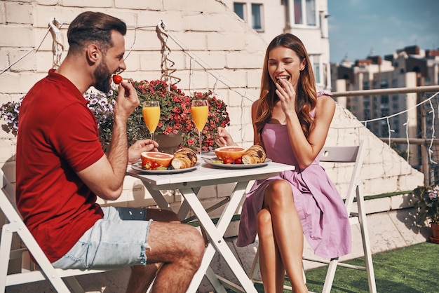 Cute young couple in casual clothing having breakfast and smiling while sitting on the rooftop patio outdoors