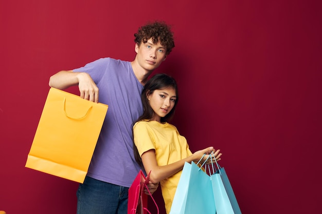 バッグとカラフルなTシャツのかわいい若いカップルショッピング赤い背景は変更されていません