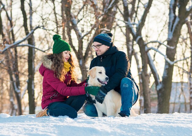 Cute young couple having fun in winter park with their dog golden retriever on a sunny day and smiling