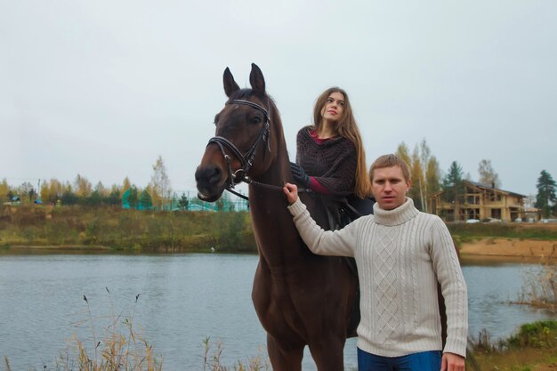 湖のそばの秋の森で馬に乗ってかわいい若いカップル。ライダーの女性と男性は、雨の厳しい曇りの天気で公園で彼女の馬を保持します。アウトドアライディング、スポーツ、レクリエーションのコンセプト。コピースペース