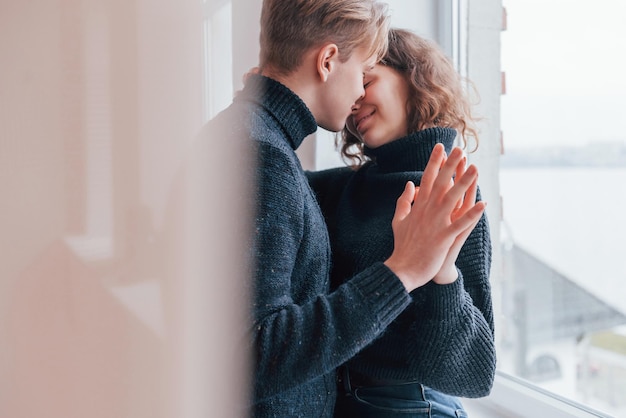 かわいい若いカップルが窓の近くで屋内でお互いにキスします。