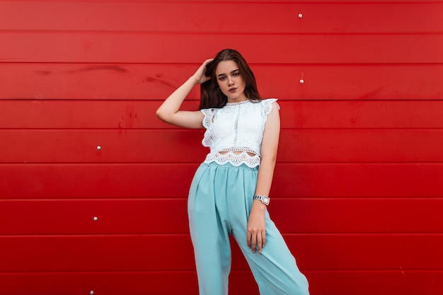 Cute young European woman in an elegant lace shirt in blue pants with brown hair stands in the city near a vintage red wooden wall.