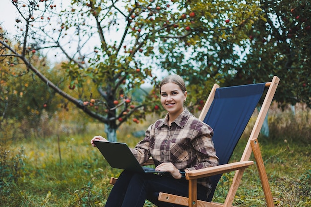 A cute young female freelancer is working in the garden outdoors while sitting in a comfortable garden chair Remote work on a portable laptop