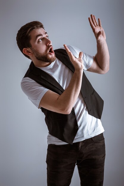 Cute young frightened man with a beard in a white shirt and a black waistcoat over gray background